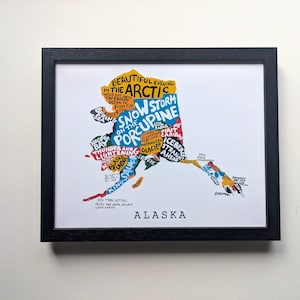 May include: A framed print of a map of Alaska, featuring colourful text-based illustrations that form the shape of the state. The text includes phrases like 'Beautiful evening in the Arctic', 'Snow storm on the Porcupine', and 'Alaska' at the bottom.
