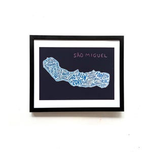 May include: A framed print featuring a dark navy background with a white outline map of São Miguel island. The map is filled with blue hand-lettered words related to the island, including The Azores, Forest, and Hiking, set in a black frame.