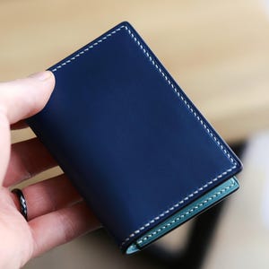 May include: A rectangular navy blue leather card holder featuring white contrast stitching along the edges. The interior of the wallet reveals a light mint green leather lining, providing a distinct color contrast against the dark blue exterior.