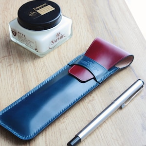 Puede incluir: Un estuche para bolígrafos de cuero azul y burdeos con un bolígrafo plateado sobre una superficie de madera. El estuche para bolígrafos tiene una solapa con el texto "Hand Made Goods" en él. También se puede ver un tarro de betún para zapatos en el fondo.