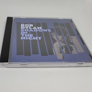 May include: A CD case with a blue background and a black spine. The front of the case features a black and white photo of Bob Dylan with the text "Bob Dylan Shadows In The Night" in white.