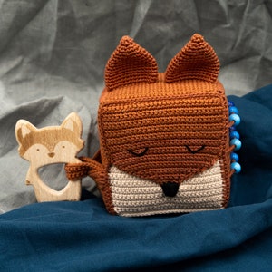 Pode incluir: Um brinquedo de crochê em forma de raposa com um rosto bege e uma pelagem marrom. O brinquedo é um cubo com contas azuis em um lado. Um mordedor de madeira em forma de raposa está ao lado do brinquedo.