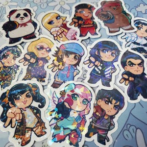 Puede incluir: Una colección de 12 pegatinas holográficas con varios personajes de dibujos animados. Las pegatinas están sobre un fondo blanco y tienen un acabado brillante e iridiscente. Los personajes incluyen un panda, un oso, una niña con cabello rubio, una niña con cabello azul, una niña con cabello rosa, un niño con cabello azul, un niño con cabello marrón y una niña con cabello negro.