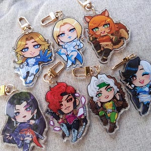 Op de afbeelding: Een verzameling van zeven acryl sleutelhangers met chibi-stijl personages met diverse haarkleuren, kostuums en poses. Elke sleutelhanger is voorzien van een goudkleurige metalen karabijnhaak aan de bovenzijde.