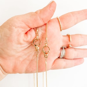 May include: Two gold-colored chain necklaces with lobster clasp closures. The necklaces are held in a hand, with several gold rings on the fingers. The chains are delicate and the clasps are detailed.