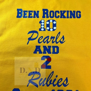 May include: Yellow t-shirt with blue text that reads "Been Rocking 10 Pearls and 2 Rubies Since 1991". The number 10 has white polka dots and the number 2 has red polka dots.