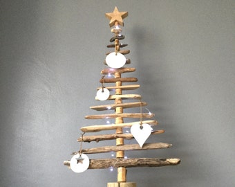 May include: A rustic wooden Christmas tree made from driftwood branches, decorated with white ceramic ornaments and a star on top. The tree is lit with white fairy lights and sits on a burlap table runner.