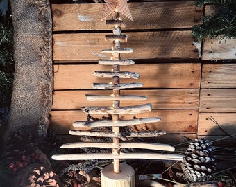 May include: A handcrafted wooden Christmas tree with a star topper. The tree is made from driftwood and small lights are strung throughout. The tree is set on a wooden base and surrounded by pine cones and greenery.