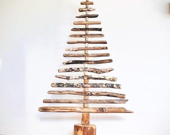 May include: A rustic wooden Christmas tree sculpture. The tree is constructed from various lengths of natural wood pieces, arranged in a triangular shape. A wooden star tops the tree, and the base is a section of a tree trunk. The tree stands on a wooden floor.