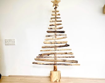 May include: A rustic wooden Christmas tree with a star topper. The tree is constructed from various pieces of driftwood arranged in a triangular shape. It stands on a wooden base and is displayed against a white wall. A unique, natural holiday decoration.