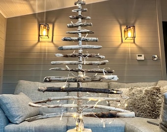 May include: A rustic driftwood Christmas tree with a star on top and white fairy lights. The tree is standing on a wooden stump and is in front of a gray couch.