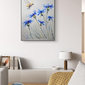 May include: Watercolor painting of blue cornflowers with a bee flying above them. The flowers are painted in shades of blue and green, with a white background.