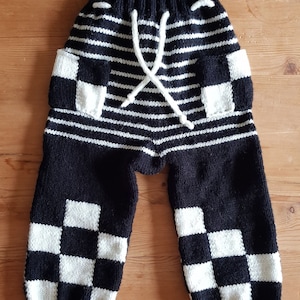 May include: Hand-knitted black and white striped pants with checkerboard accents. The pants feature a drawstring waist and checkerboard pockets and cuffs. The color scheme is black and white.