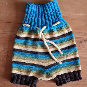 May include: Hand-knitted baby shorts with a blue ribbed waistband and a drawstring. The shorts feature horizontal stripes in shades of blue, green, cream, and dark gray. The bottom hem is dark gray.