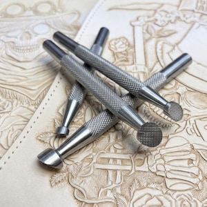 May include: A set of four silver-colored metal leather stamping tools with knurled handles and textured circular heads, resting on a piece of light tan leather featuring an embossed design of a skull wearing a top hat and roses.