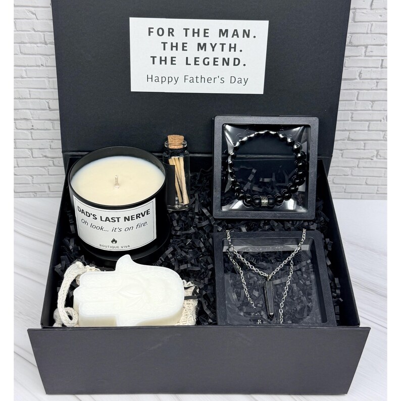 Op de afbeelding: Een zwarte geschenkdoos met een witte kaars met het label DAD'S LAST NERVE Oh look... it's on fire, een klein glazen flesje met lucifers, een zwarte kralenarmband, een zilverkleurige ketting met een donkere hanger en een witte hamsa-zeep.