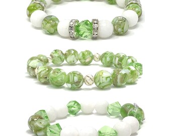 May include: Three green and white beaded bracelets with silver accents. The top bracelet has clear crystal accents. The middle bracelet has a silver charm. The bottom bracelet has white beads.