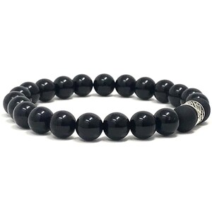 Op de afbeelding: Een elastische armband bestaande uit gladde, ronde, gepolijste zwarte kralen. De armband is voorzien van een enkele zilverkleurige metalen tussenkraal met een ingewikkeld, gegraveerd patroon, geplaatst nabij een uiteinde van de cirkelvormige opstelling.