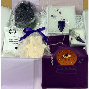 May include: A gift box filled with spiritual items. Includes a large amethyst geode, a necklace with a purple pendant, a pendulum, a purple velvet bag with a crystal ball design, and a small package with the text "A LITTLE EXTRA LOVE".