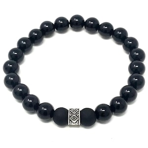 Op de afbeelding: Een ronde elastische armband bestaande uit gladde, glanzende zwarte kralen. Het ontwerp bevat twee matte zwarte kralen die een centrale zilverkleurige cilindrische tussenkraal flankeren, versierd met een ingewikkeld gegraveerd geometrisch patroon.