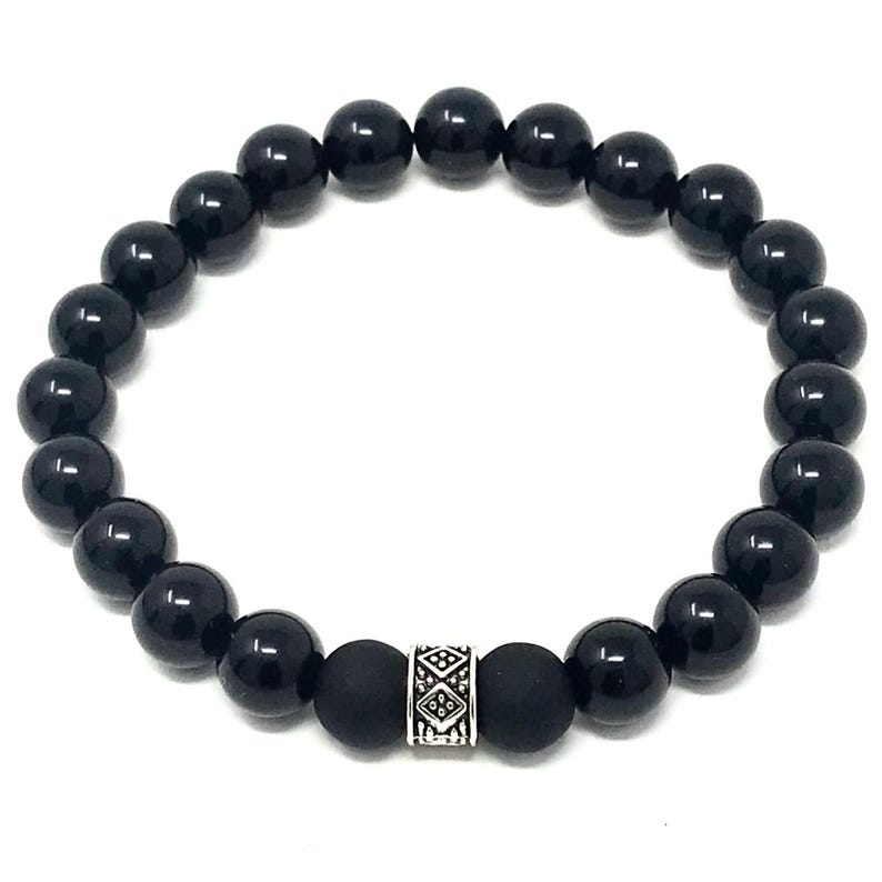 Op de afbeelding: Een ronde elastische armband bestaande uit gladde, glanzende zwarte kralen. Het ontwerp bevat twee matte zwarte kralen die een centrale zilverkleurige cilindrische tussenkraal flankeren, versierd met een ingewikkeld gegraveerd geometrisch patroon.