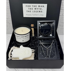 Op de afbeelding: Een zwarte geschenkdoos met een witte kaars met het label DAD'S LAST NERVE Oh look... it's on fire, een klein glazen flesje met lucifers, een zwarte kralenarmband, een zilverkleurige ketting met een donkere hanger en een witte hamsa-vormige zeep.