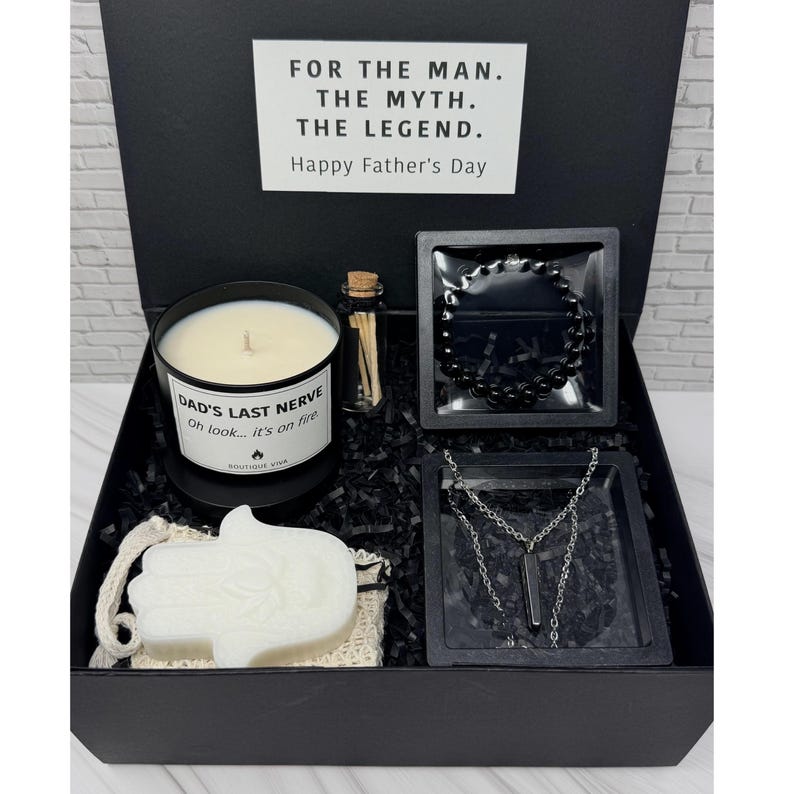 Op de afbeelding: Een zwarte geschenkdoos met een witte kaars met het label DAD'S LAST NERVE Oh look... it's on fire, een klein glazen flesje met lucifers, een zwarte kralenarmband, een zilverkleurige ketting met een donkere hanger en een witte hamsa-vormige zeep.
