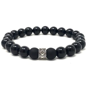 Op de afbeelding: Een elastische armband bestaande uit gladde, gepolijste zwarte kralen en twee matte zwarte kralen die een centrale zilverkleurige metalen kraal flankeren met een ingegraveerd geometrisch patroon op het oppervlak.