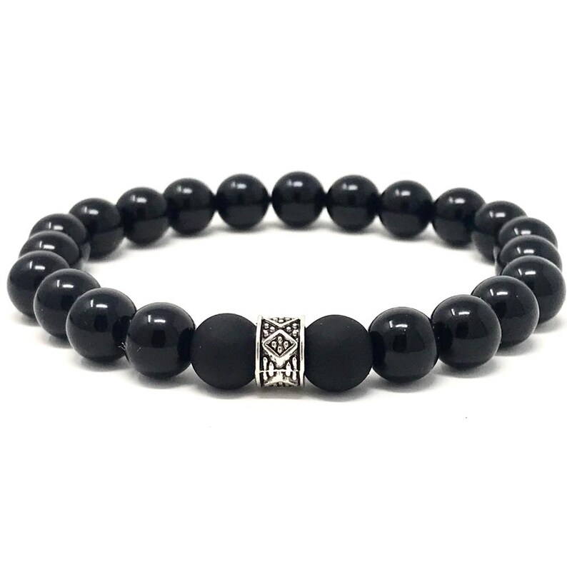 Op de afbeelding: Een elastische armband bestaande uit gladde, gepolijste zwarte kralen en twee matte zwarte kralen die een centrale zilverkleurige metalen kraal flankeren met een ingegraveerd geometrisch patroon op het oppervlak.