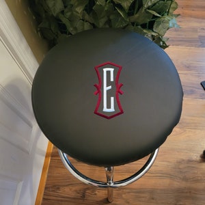 Puede incluir: Un taburete de bar negro con un asiento redondo de cuero negro. El asiento tiene una letra "E" bordada en rojo en el centro.