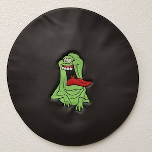 以下が含まれることがあります： 黒い丸い布のパッチに、赤い舌を出した緑色の漫画の幽霊が描かれています。幽霊はパッチに刺繍されています。