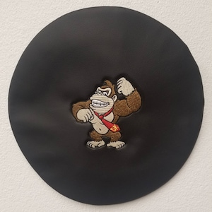 May include: Black beret featuring an embroidered image of Donkey Kong flexing his muscles. The character is brown with a red tie and the initials "DK" on it. The beret is round and made of a smooth, dark material.
