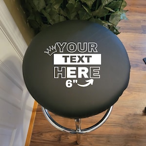 May include: Black bar stool seat cover with a white crown and the text "YOUR TEXT HERE 6" printed on it.
