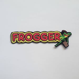 Könnte beinhalten: Ein gestickter Aufnäher mit dem Wort "FROGGER" in roter und gelber Schrift. Ein grüner Froschcharakter springt rechts neben dem Wort.