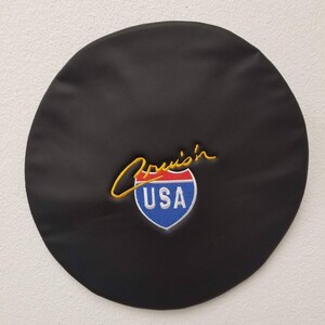 May include: Black leather car steering wheel cover with gold embroidery that reads "Cruisin' USA" and a red, white, and blue highway sign.