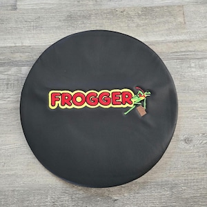 May include: A black, round cover featuring the word "FROGGER" in yellow and red lettering. A green frog character with a briefcase is embroidered on the cover. The cover is made of a smooth, dark material.