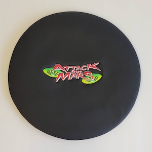 May include: A round, black cushion with the embroidered text "ATTACK FROM MARS" in red, white, and green. The cushion is circular and appears to be made of a smooth, synthetic material.