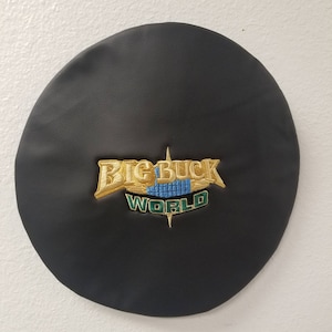 May include: A black, round, leather-like beret with an embroidered "BIG BUCK WORLD" logo in gold, blue, and green. The logo is centered on the beret, which is against a white background.