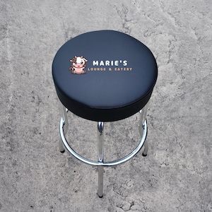 May include: Black bar stool with a chrome base and a black padded seat. The seat has a logo that says "Marie's Lounge & Eatery" with a cartoon cow.