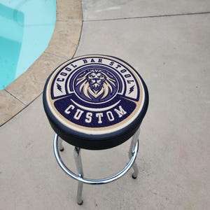 May include: A black bar stool with a round, black leather seat. The seat has a custom embroidered design of a lion's head with the words "Cool Bar Stool Custom" around it.