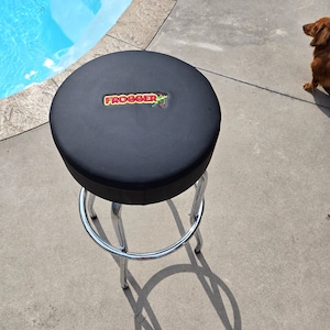 May include: Black bar stool with a round, black leather seat. The seat has a red and yellow embroidered logo that says "FROGGER" with a green frog graphic.