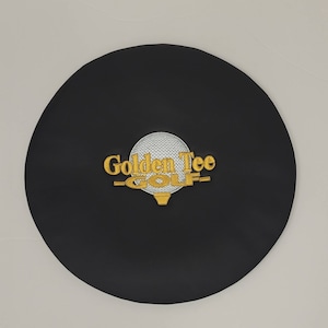 May include: A black, circular golf club head cover with the words "Golden Tee GOLF" embroidered in gold thread. The golf ball graphic is white with a gold tee.