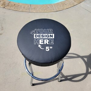 May include: A black, round bar stool with a chrome base. The seat has the text "YOUR DESIGN HERE" and an arrow pointing to "5". The stool is outdoors near a pool.