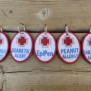 Può includere: Cinque portachiavi ovali bianchi con bordi rossi e una croce rossa nell'angolo in alto a sinistra. Ogni portachiavi ha un diverso messaggio di allerta medica: Allerta Asma, Allerta Diabete, EpiPen, Allergia alle Arachidi e Allergia ai Frutti di Mare.