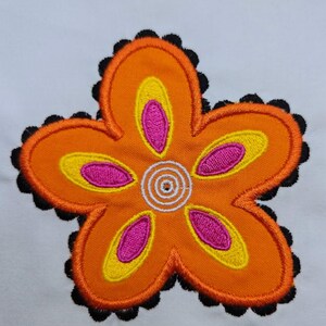 May include: An embroidered flower applique with orange petals, yellow and pink accents, and a black scalloped border. The center features a white spiral design. This floral design is likely for crafts or fabric embellishment.