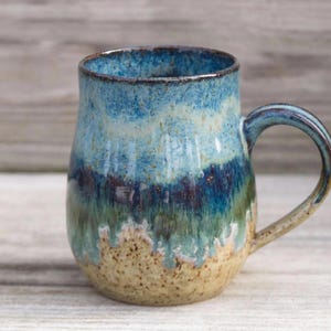 May include: A handmade ceramic mug with a unique glaze. The mug features a gradient of colors, including shades of blue, green, and tan. The handle is a matching blue. The mug is designed for hot beverages.