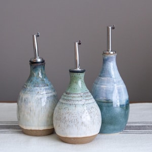 May include: Three ceramic oil dispensers with rounded bodies and narrow necks. Each bottle features a unique glaze in shades of blue, green, and white, with a visible brown clay base. Each dispenser is topped with a silver metal pour spout.