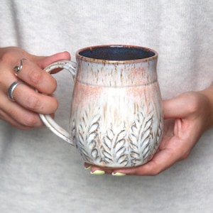 May include: A ceramic mug with a rounded body and a handle, featuring a white and light pink glaze with a textured leaf pattern embossed around the base. The interior of the mug is a dark blue colour, and the rim has a thin brown edge.