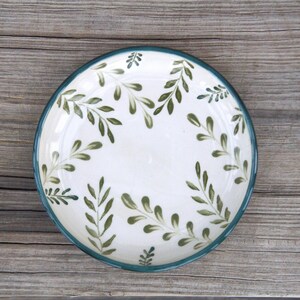 May include: A round ceramic plate with a white base and a hand-painted pattern of green leafy branches. The plate features a dark green rim that frames the botanical design, which is distributed evenly across the surface of the dish.