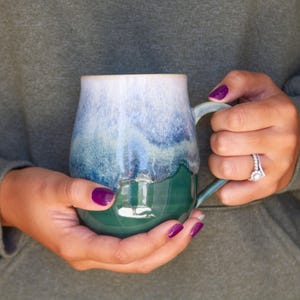 May include: A handmade ceramic mug with a unique glaze. The mug features a gradient of colors, transitioning from white at the rim to shades of blue and green at the base. The mug is held by a person wearing a green hoodie.